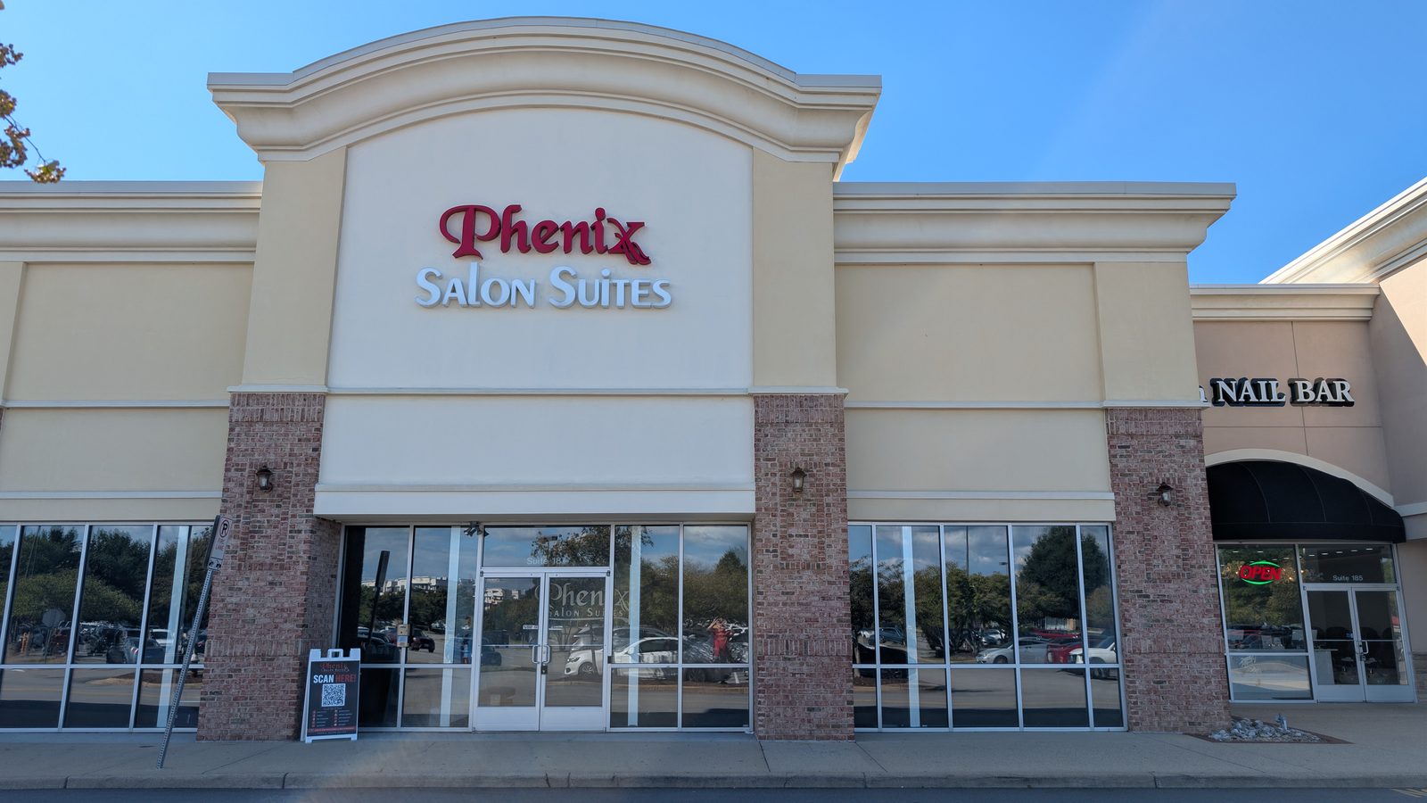 The Phenix Salon Suites storefront seen from the parking lot: a cream building with a raised arched parapet, the Phenix Salon Suites sign in red and grey lettering, and glass double doors between brick pillars. A nail bar occupies the unit to the right.