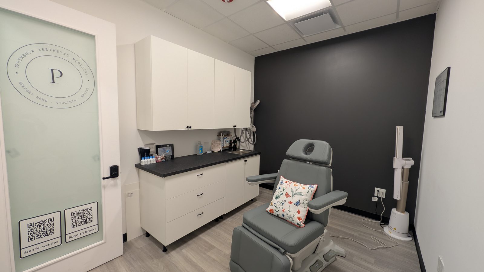 One of the two treatment rooms: a grey powered treatment chair with a face cradle, a run of white cabinets over a dark counter with a sink, a charcoal feature wall, and a frosted glass door carrying the Peninsula Aesthetic Medicine monogram.